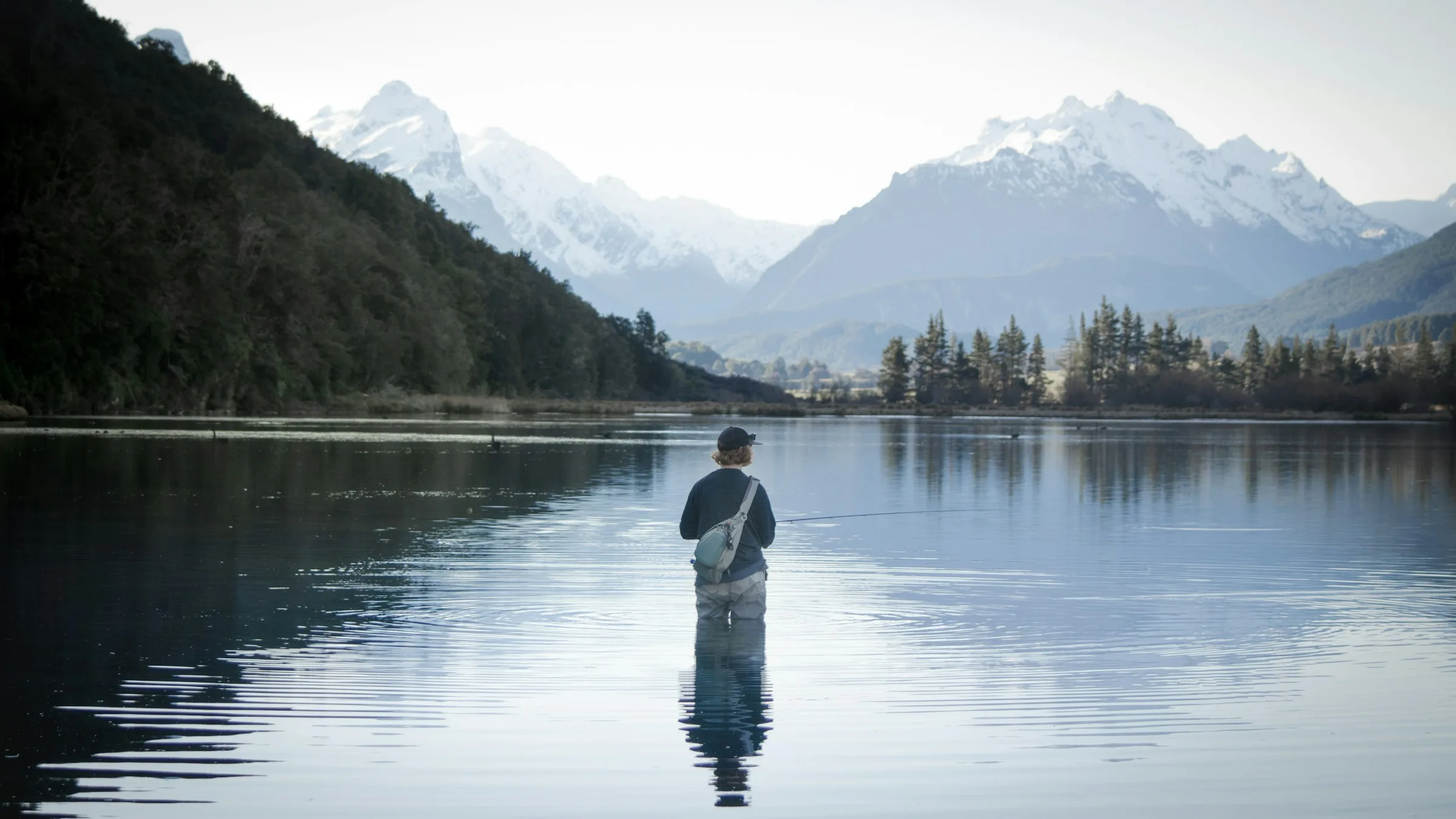 A fly angler standing in a still mountain lake at dawn, snow-capped peaks behind — New Zealand