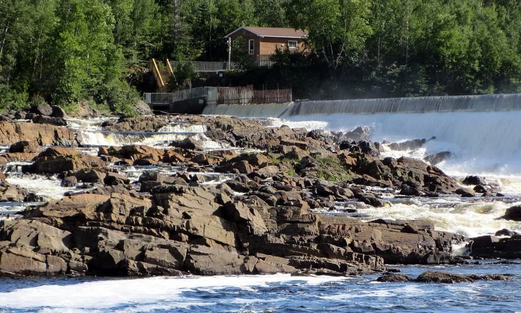 Whitewater cascading over dark rock ledges on the Exploits River, with a small dam structure and building among trees in the background.