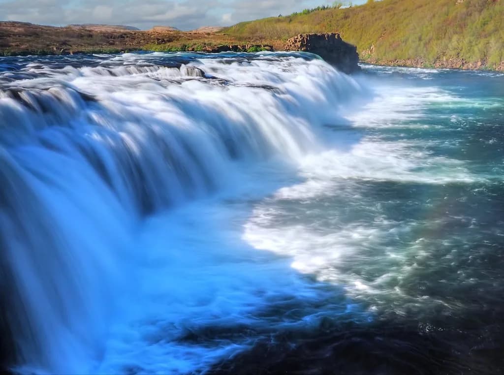 The Faxi waterfall cascading over a wide rock ledge on the Tungufljót River, Iceland, with blue-white whitewater and a grassy far bank.