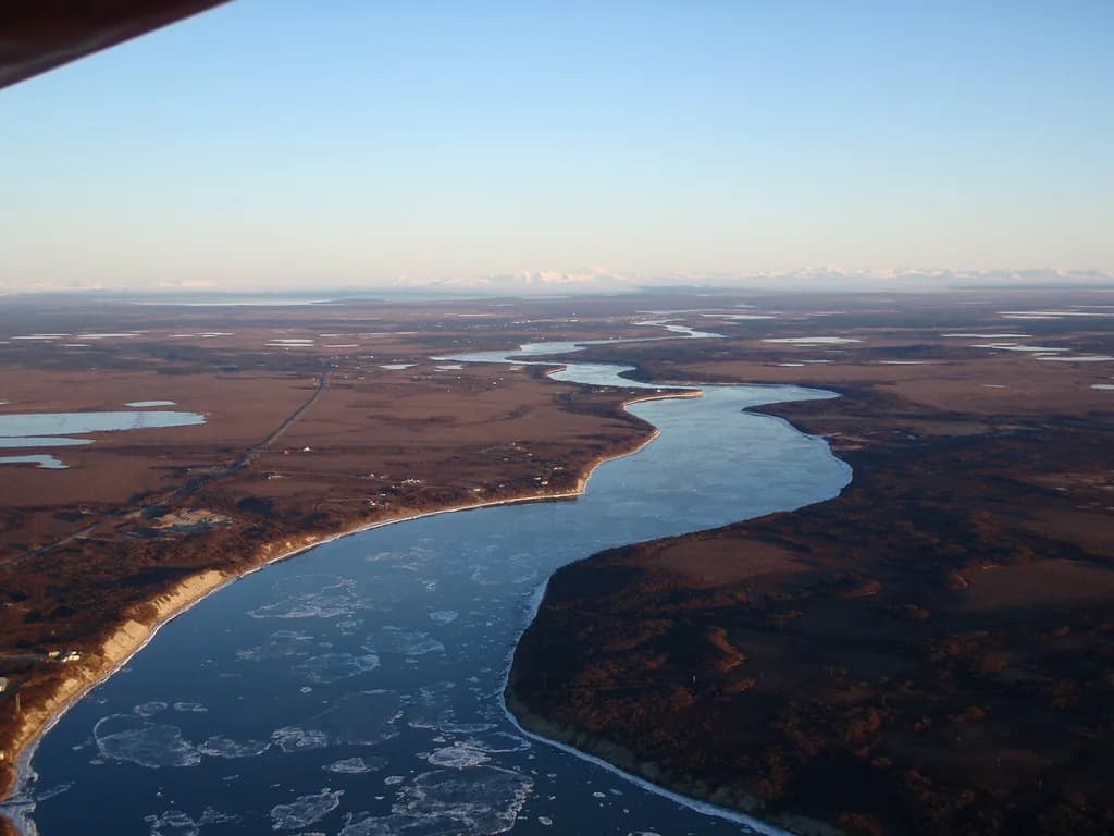Aerial view of the Naknek River winding through tundra wetlands toward Bristol Bay, Alaska, with mountains on the horizon.