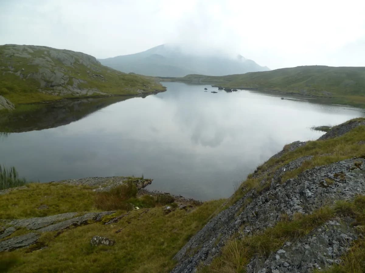 Calm upland lake (Llyn Cwm Corsiog) surrounded by rocky, grassy banks with misty hills in the background, Snowdonia.