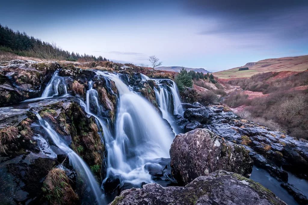 A wide waterfall cascading over dark rock ledges on the River Endrick at the Loup of Fintry, with moorland hills in the background.