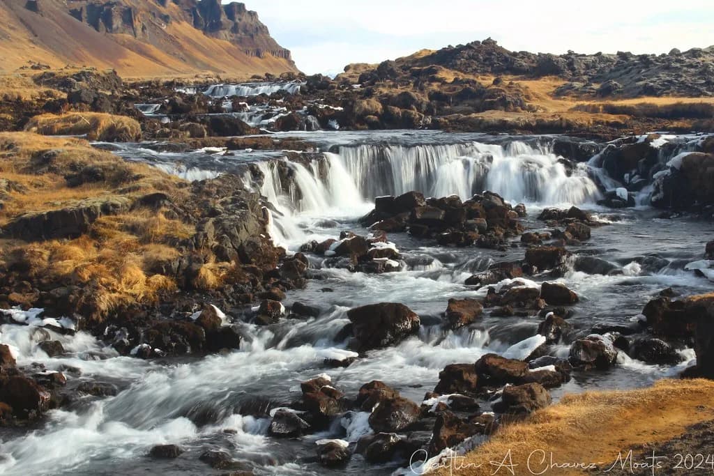 Multiple cascading waterfalls of the Fossalár river flowing over dark volcanic rock in Iceland, with golden autumn grass and mountains in the background.