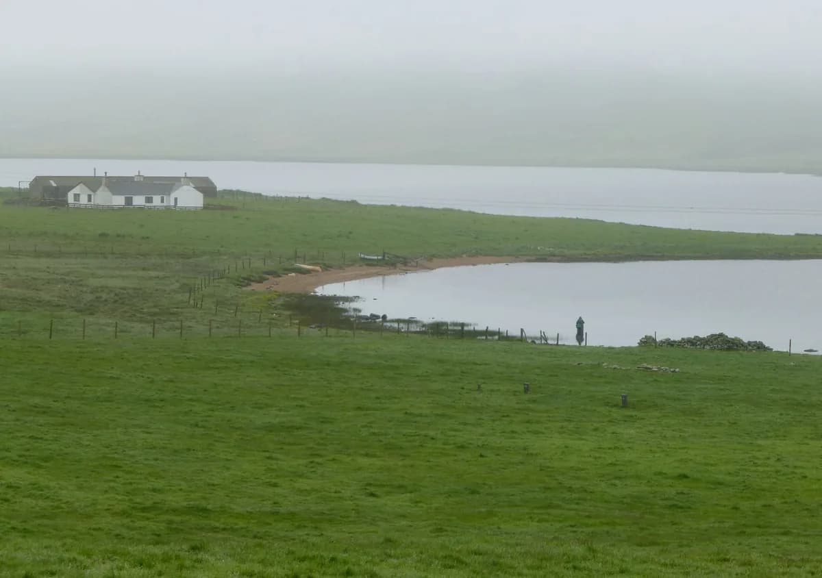 A lone fisherman casts from the shore of the Loch of Spiggie in Shetland, with open water and low hills in the background.