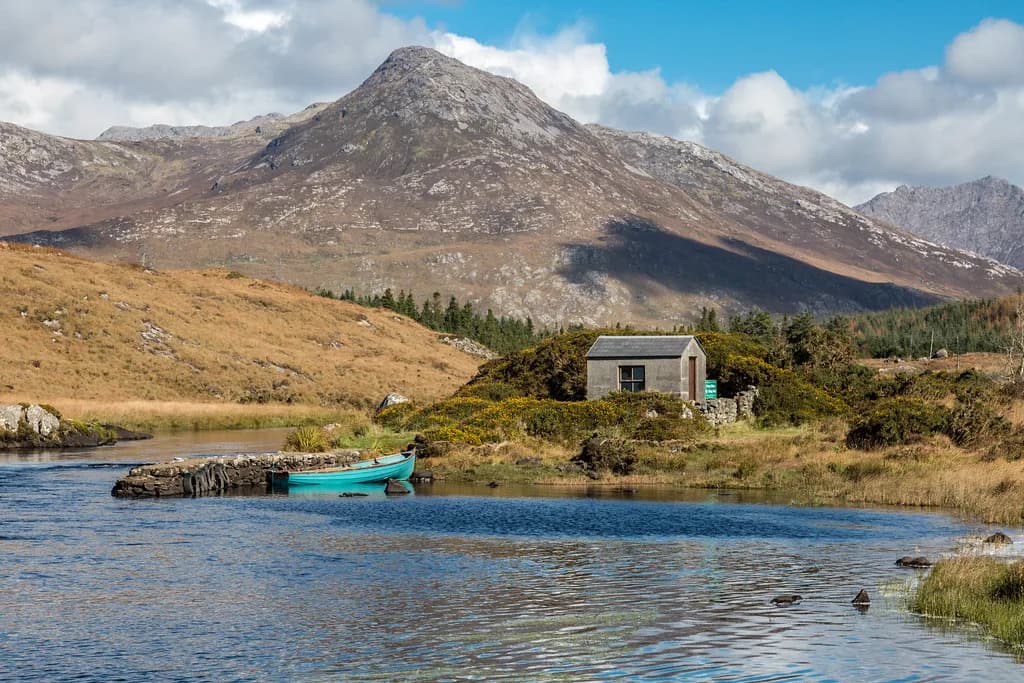 A turquoise fishing boat tied up beside a stone boathouse on the water at Ballynahinch, with mountains in the background.