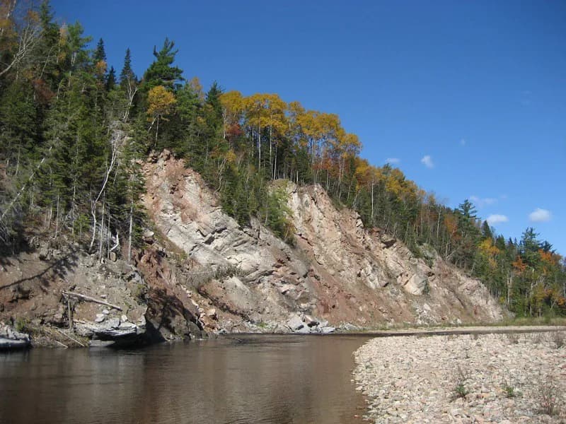 The Margaree River bending past a steep, layered rock cliff and autumn-coloured trees, with a gravel bar in the foreground.