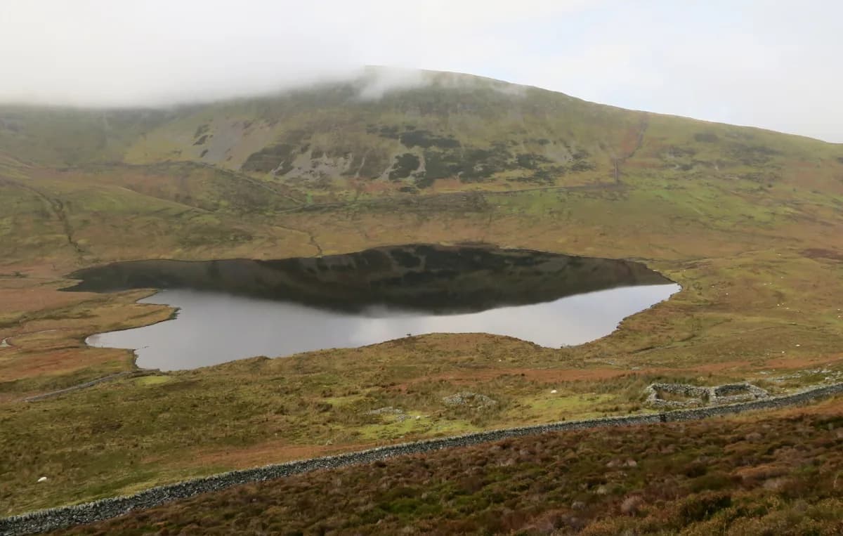 A dark, still upland lake filling a grassy hollow, with mist clinging to the hillside above and a stone wall in the foreground.