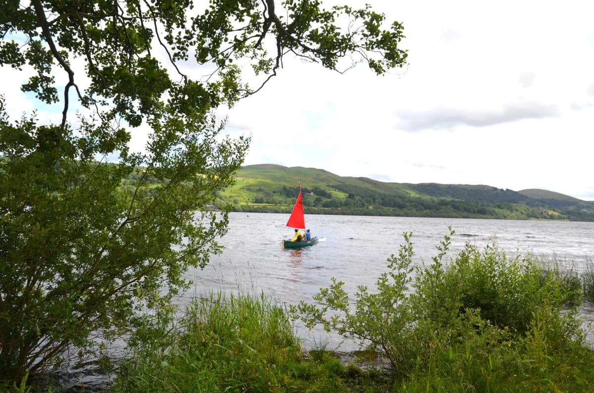 Wide view of Bala Lake (Llyn Tegid) from the Llanycil shoreline, with a small sailing boat on the water and hills in the background, framed by lakeside trees.