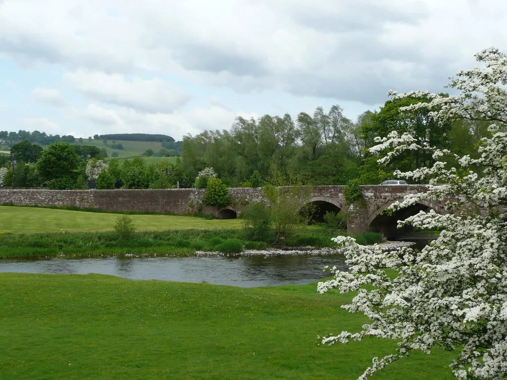A calm stretch of the River Eamont in Cumbria, with a stone arched bridge, hawthorn blossom, and green meadows.