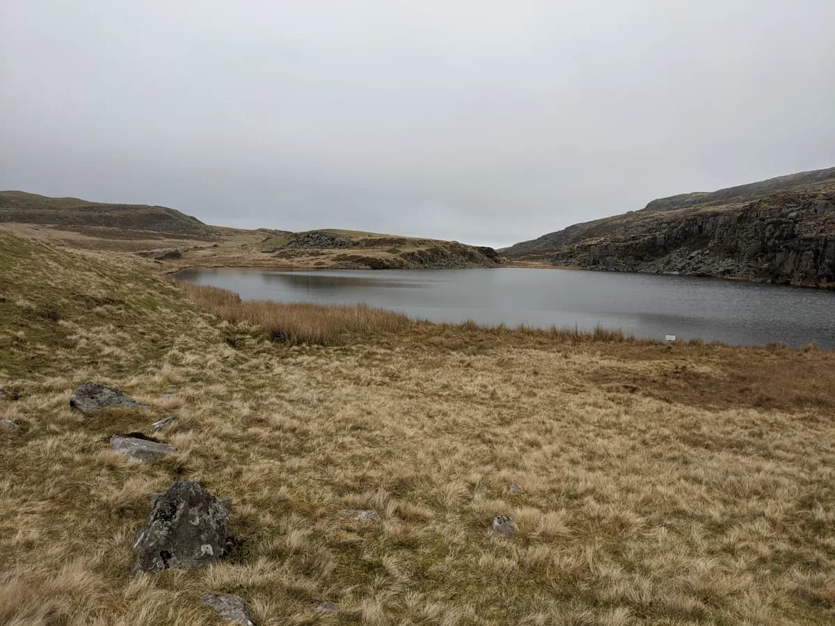 A grey, still upland lake bordered by tussocky moorland grass and a low rocky outcrop, under an overcast sky.