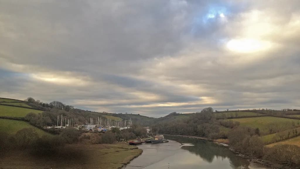 A wide view of the River Lynher valley in Cornwall, with moored boats and rolling green hills under a cloudy sky.