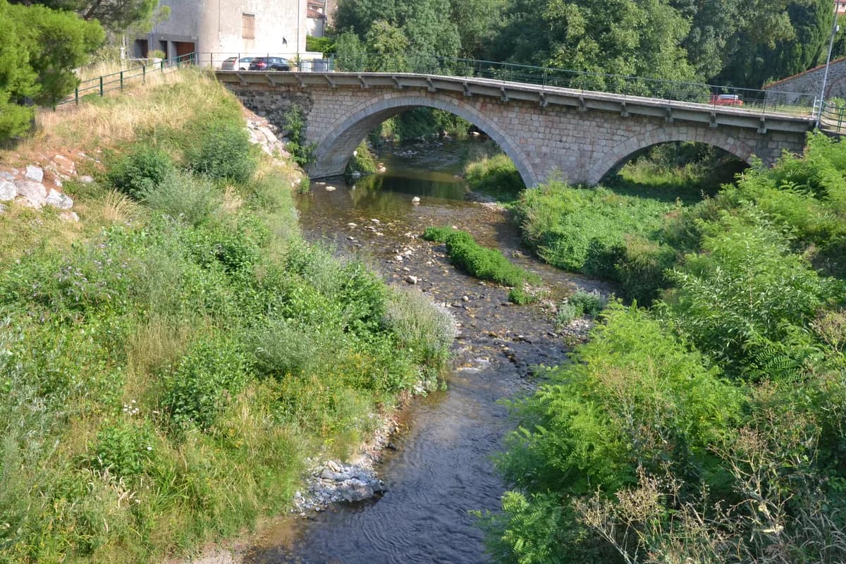 A stone arch bridge crossing the shallow, rocky Boulzane river lined with dense green vegetation in the Fenouillèdes region of France