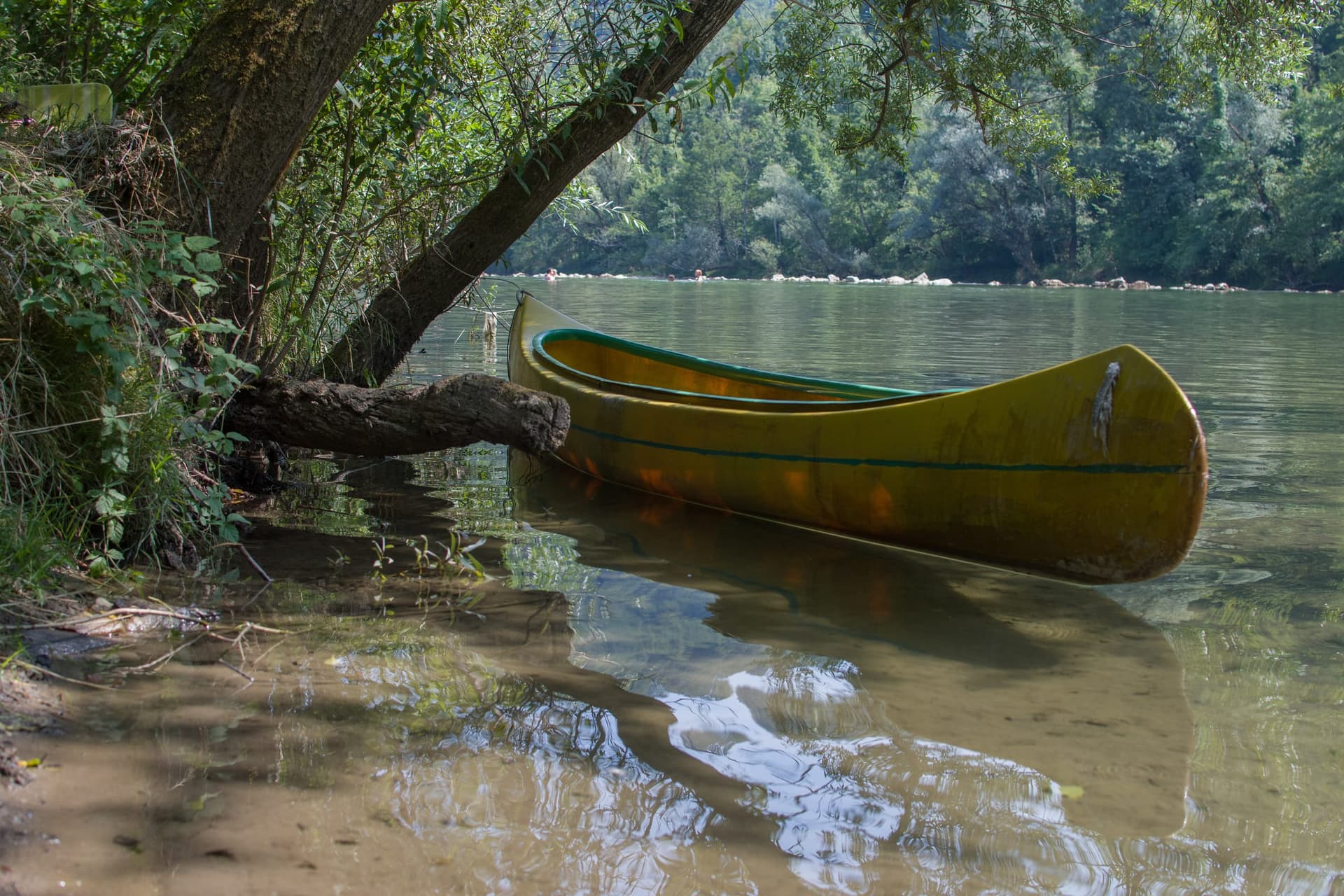 A canoe resting on the calm green bank of the Kolpa beneath overhanging willows, forested hills behind.