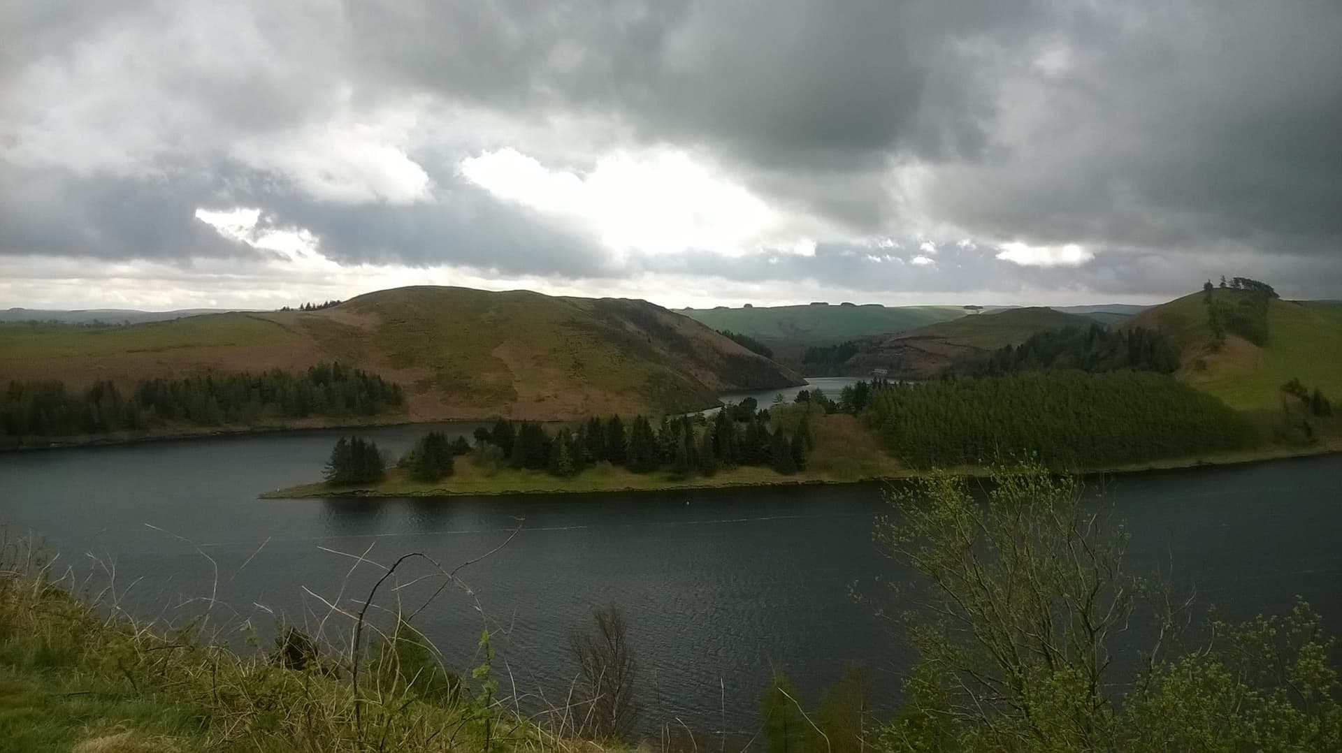Open water of Llyn Clywedog reservoir from the shoreline, hills and sky reflected in the calm surface.