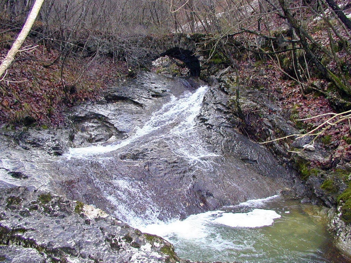 A clear stream cascading over limestone slabs beneath a stone arch bridge in the Srna gorge, Gorenja Trebuša.